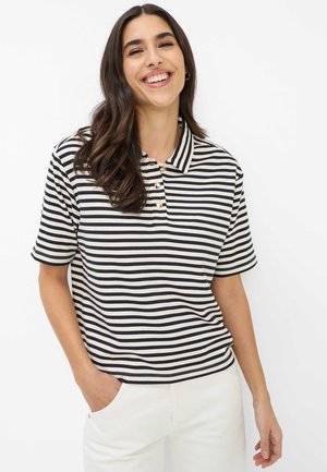 Junge Frau, die lächelt, trägt ein schwarz-weiß gestreiftes Polohemd mit kurzen Ärmeln und eine weiße Hose, mit der Hand in der Tasche.