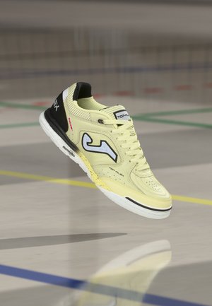 Gelber und schwarzer Hallensportschuh mit weißer Sohle, Markenlogo an der Seite, geschnürte Schnürsenkel, schwebend über einem polierten Hallenboden mit farbigen Linien.