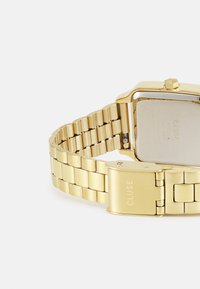 Montre-bracelet en métal doré avec un cadran rectangulaire, bracelet à maillons et fermoir. Le dos présente la gravure de la marque et des spécifications.