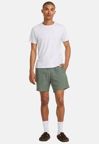 T-shirt en coton blanc, short vert avec des rayures jaunes verticales, poches latérales, porté avec des chaussettes blanches et des chaussures à enfiler marron.