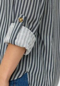 Manche de chemise à rayures roulée avec un rabat boutonné, portée par une personne, montrant des rayures verticales bleu marine et blanches ainsi qu'une partie de jean bleu.