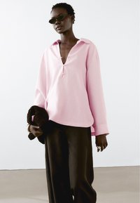 Růžová košile s dlouhým rukávem a límečkem, se dvěma knoflíky a volným střihem. Modelka drží tmavý pletený doplněk.