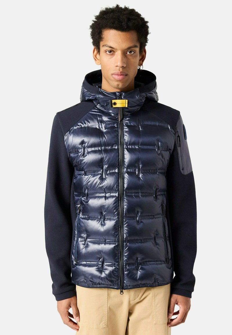 Granatowa kamizelka puchowa o błyszczącej pikowanej powierzchni, z przypinaną kapturem i żółtym zapięciem na zatrzask. Długie rękawy z kontrastowego materiału.