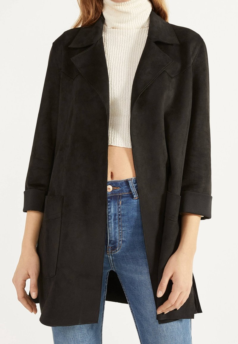 Donna che indossa un cappotto nero aperto con maniche arrotolate sopra un top corto a collo alto bianco a coste e jeans blu a vita alta.