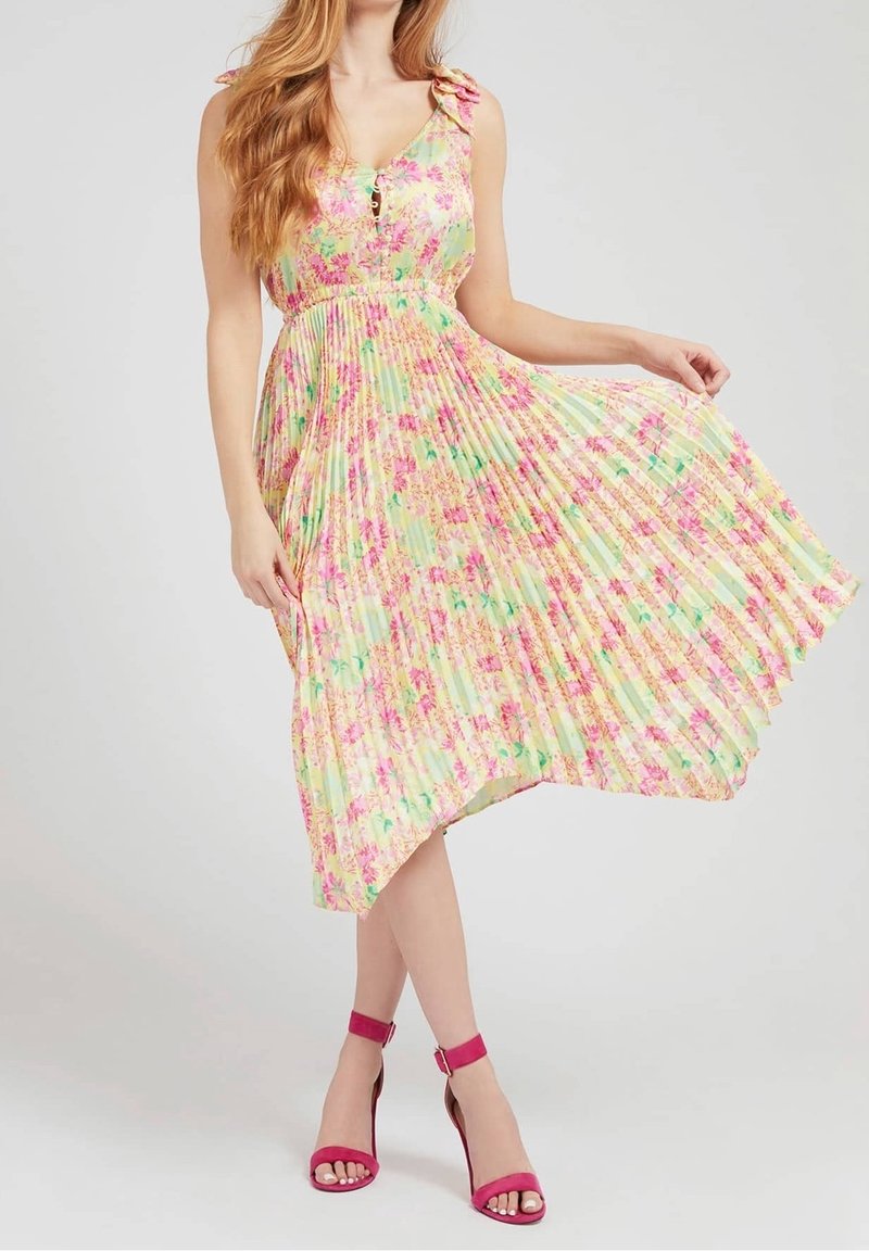 Femme en robe plissée fleurie pastel sans manches, tenant sa jupe, portant des talons à bride à la cheville fuchsia, posant sur fond gris uni.