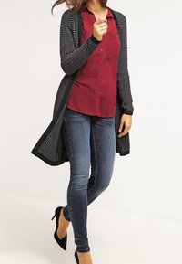 Long black cardigan with textured knit, worn over a burgundy button-up shirt, paired with dark blue skinny jeans and black heels.
