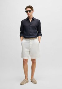 Completo maschile con una camicia button-up blu scuro, pantaloni corti bianchi e scarpe slip-on beige con texture intrecciata. Include occhiali da sole e una cintura.