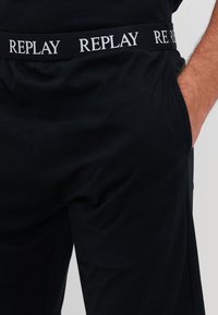 Pantalones jogger negros con una textura suave y una cintura elástica con el logotipo "REPLAY" en blanco repetido. Bolsillos laterales visibles.