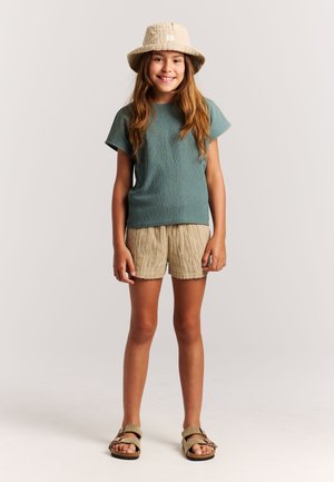 Turquoise textuur t-shirt met korte mouwen, beige textuurs korte broek en beige bucket hat. Bruine sandalen met open tenen en bandjes. Eenvoudige, casual outfit.