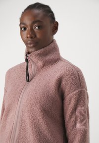 Fleece jacket in soft pink with a high collar, full zip, and textured fabric. Features a logo patch on the left arm.
