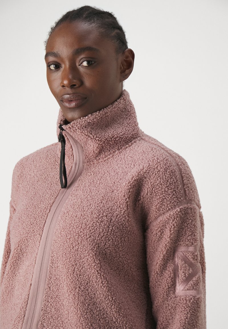 Fleece-Jacke in zartem Rosa mit hohem Kragen, durchgehendem Reißverschluss und strukturiertem Stoff. Mit einem Logo-Patch am linken Arm.