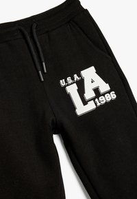 Pantalones deportivos negros con cintura elástica, que presentan un gráfico blanco que dice "U.S.A. LA 1986" en el lateral y un bolsillo lateral.