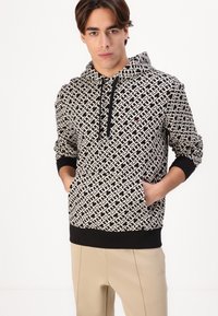 Young man wearing a black-and-white patterned hoodie with front pocket and beige pants, standing against a plain white background.