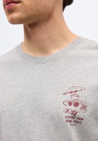 Άνδρας με ανοιχτόγριλο πουκάμισο με μικρό γραφικό σχεδιο γυαλιών ηλίου, ποτού και χάμπουργκερ κάτω από ομπρέλα, κείμενο «Κρατώντας τα ψωμάκια ζεστά.»