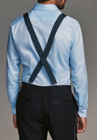 Dark blue suspenders attached to a light blue shirt, intersecting at the back, with silver hardware and a textured fabric. Dark trousers visible.