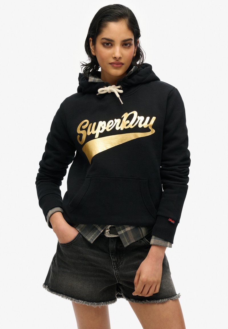 Sudadera negra con el logo dorado de "SuperDry", con capucha ajustable, bolsillo frontal y puños acanalados. Combinada con pantalones cortos de mezclilla grises.