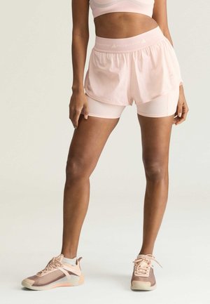 Kvinde iført lyserøde lagdelte træningsshorts, matchende sports-bh og beige-lyserøde sneakers, stående mod en ensfarvet baggrund.