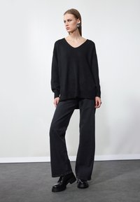 Un pull noir oversize avec un col en V, associé à un jean noir à large jambe. Le mannequin porte des bottes plateformes noires épaisses et de grandes boucles d'oreilles.