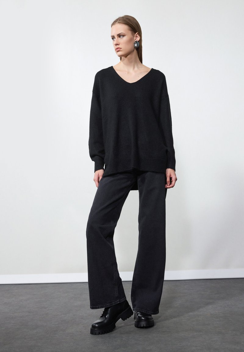 Čierny oversized sveter s V-výstrihom, skombinovaný s voľnými čiernymi džínsami. Model nosí hrubé čierne platformové topánky a veľké náušnice.