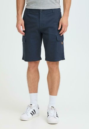 Mørk marineblå cargoshorts laget av bomull, med to sidelommer, rett snitt og knappelukking. Brukt med hvite sokker og joggesko.