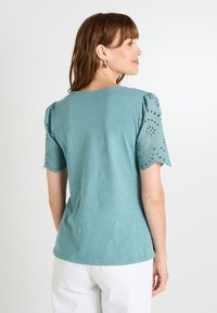 Top corto de color verde azulado con detalles de ojales en las mangas, escote redondo y un corte relajado, combinado con pantalones blancos. Tejido suave y ligero.