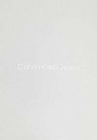 Vitt stretchtyg med ribbad textur som har "Calvin Klein Jeans"-logotypen tydligt centrerad i en djärv, modern stil.