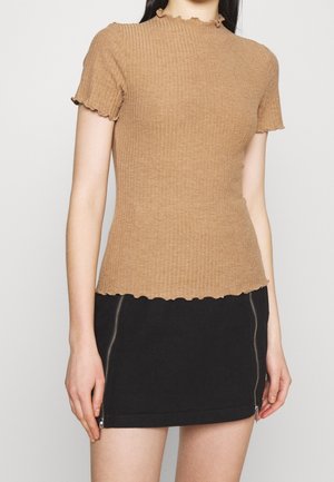 Beżowa prążkowana bluzka z krótkimi rękawami i falbankowanym dekoltem; noszona z czarną mini spódniczką z bocznym zamkiem błyskawicznym. Widoczna faktura materiału.