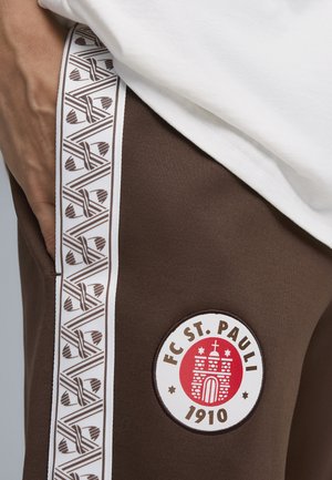 Brune atletiske bukser med en hvid sidebånd med et geometrisk mønster og et cirkulært broderet logo for FC St. Pauli, grundlagt i 1910.