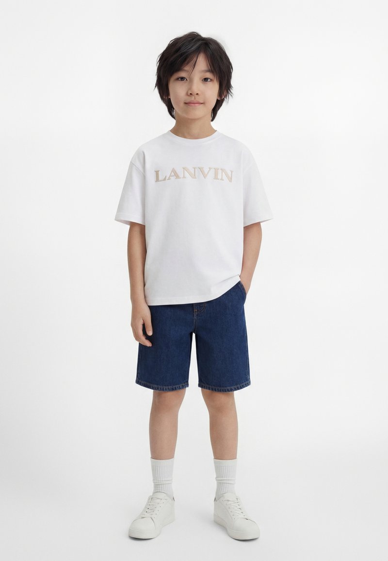 Garçon aux cheveux noirs se tenant face à nous, portant un t-shirt blanc Lanvin, un short en jean bleu foncé, des chaussettes blanches et des baskets blanches.