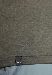 Olivengroene T-shirt met een gladde textuur, voorzien van een zwart label aan de zoom met een wit logo, gecombineerd met een lichtblauwe stof eronder.
