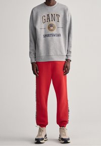 Šedá mikina s potiskem "GANT SPORTSWEAR", spárovaná s červenými teplákovými kalhotami s bočními pruhy. Model má na sobě béžové tenisky.