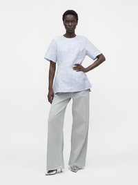 Femme debout, une main sur la taille, portant un haut bleu clair à motifs à manches courtes, un pantalon large gris pâle et des talons pointus argentés.
