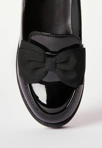 Czarny lakierowany but ze skórzaną, zaokrągloną na przodzie częścią oraz czarną, teksturowaną kokardą z przodu. Gładka i błyszcząca powierzchnia.