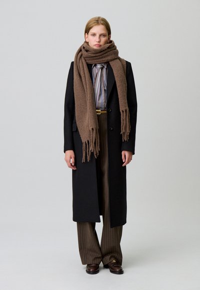 Lungo cappotto nero su una camicia a righe, sciarpa marrone e pantaloni a righe sottili. La sciarpa ha dettagli frangiati; le scarpe sono di un marrone scuro.