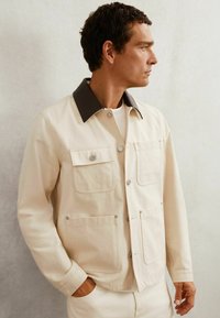 Lys beige jakke med en mørkebrun krave, der har trykknapper og dobbelte brystlommer. Fremstillet af tekstureret stof med et minimalistisk design.