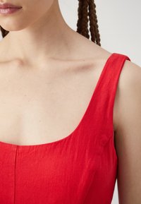 Robe en lin rouge avec un large décolleté en U et de fines bretelles, présentant une finition texturée et des coutures nettes visibles.