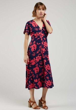 Femme portant une robe midi bleu marine à imprimé floral rouge vif et manches courtes, associée à des sandales à talons marron, debout avec la main près du visage.