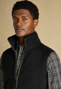 Melna flīsa veste ar augstu apkakli un zelta rāvējslēdzēju, uzlikta virs kvadrātveida krekla ar pogām, kas satur sarkanas, zaļas un zilas tonus.