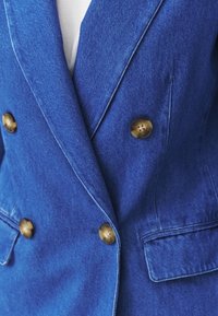 Gros plan d'une veste en denim bleue à double boutonnage avec des boutons marron et des poches avant visibles, portée sur un haut blanc.
