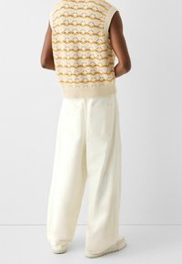 Person klädd i en krämfärgad och gul mönstrad ärmlös stickad topp med vida krämfärgade högmidjade byxor och krämfärgade sneakers, vänd bort.