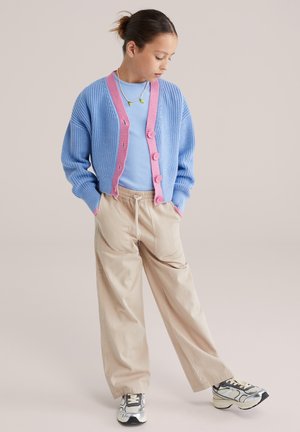 Svetlomodro pleten kardigan z rožnato obrobo in gumbi, nošen preko svetlomodrega topa in bež širokih hlač. Doplnjeno s sivimi supergami.