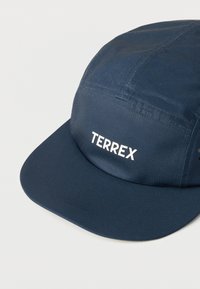 Καπέλο από ύφασμα ναυτικού μπλε με επίπεδη γείσα και λευκό λογότυπο "TERREX" στο κέντρο του μπροστινού πάνελ, σε απλό ανοιχτό φόντο.