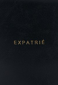 Czarna skórzana powierzchnia z wytłoczonym napisem "EXPATRIÉ" w kolorze złotym. Tekstura wydaje się gładka z lekkim wzorem słojów.