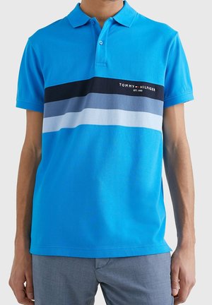 Man draagt een blauw Tommy Hilfiger polo-shirt met horizontale zwarte, grijze en witte strepen over de borst, gecombineerd met een grijze broek.