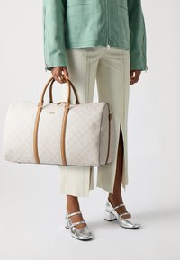 Geruite beige weekendtas met tan leren handvatten, met een ronde vorm en ritssluiting, gecombineerd met lichtgroene broeken.