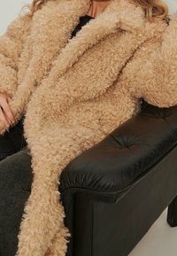 Henkilö, joka on pukeutunut beigekuituiseen tekoturkisnahkatakkiin, istuu mustassa nahkaisessa napitetussa nojatuolissa, josta näkyy tummat housut.