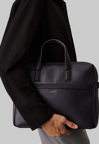 Black leather briefcase with textured surface, dual top handles, and a sleek design. Features a logo and contrasting smooth leather trim.