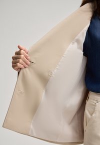 Beige blazer met een gestructureerde buitenstof en een gladde voering, voorzien van een doorzichtige knop en zichtbare stikdetails.
