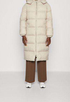 Personne portant un long manteau matelassé beige avec capuche, un pantalon large marron et des baskets blanches, debout devant un fond uni.