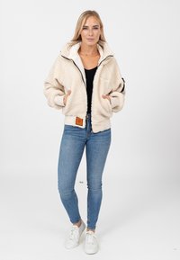 Kremowa, przytulna kurtka sherpa z kapturem, z zamkiem błyskawicznym z przodu, bocznymi kieszeniami i elastycznymi mankietami, noszona na czarnym topie i niebieskich dżinsach. Białe sneakersy.